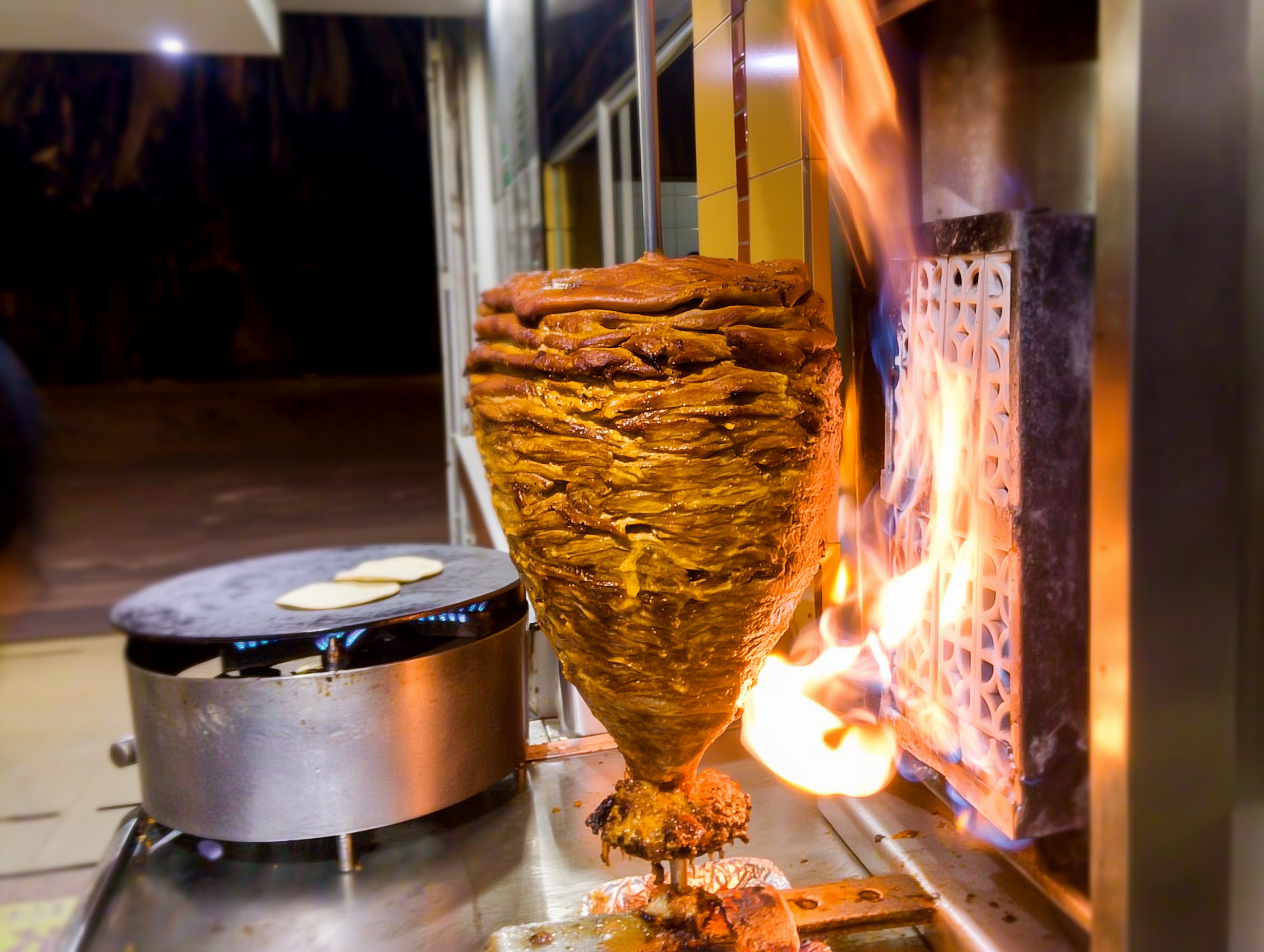Pork stacked on a trompo beside the flame, being shaved onto a tortilla.
