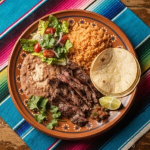 Sliced grilled steak with Mexican rice, refried beans, salad and warm tortillas, on a terracotta clay plate.