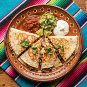 A quesadilla cut into wedges showing melted cheese and grilled meat, with salsa, guacamole and sour cream, on a terracotta clay plate.