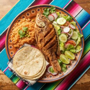 A whole crisp fried tilapia with Mexican rice, salad, lime and warm tortillas, on a terracotta clay plate.