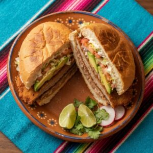A pressed torta on a telera roll cut in half, filled with a breaded steak cutlet, beans, avocado, lettuce and tomato.