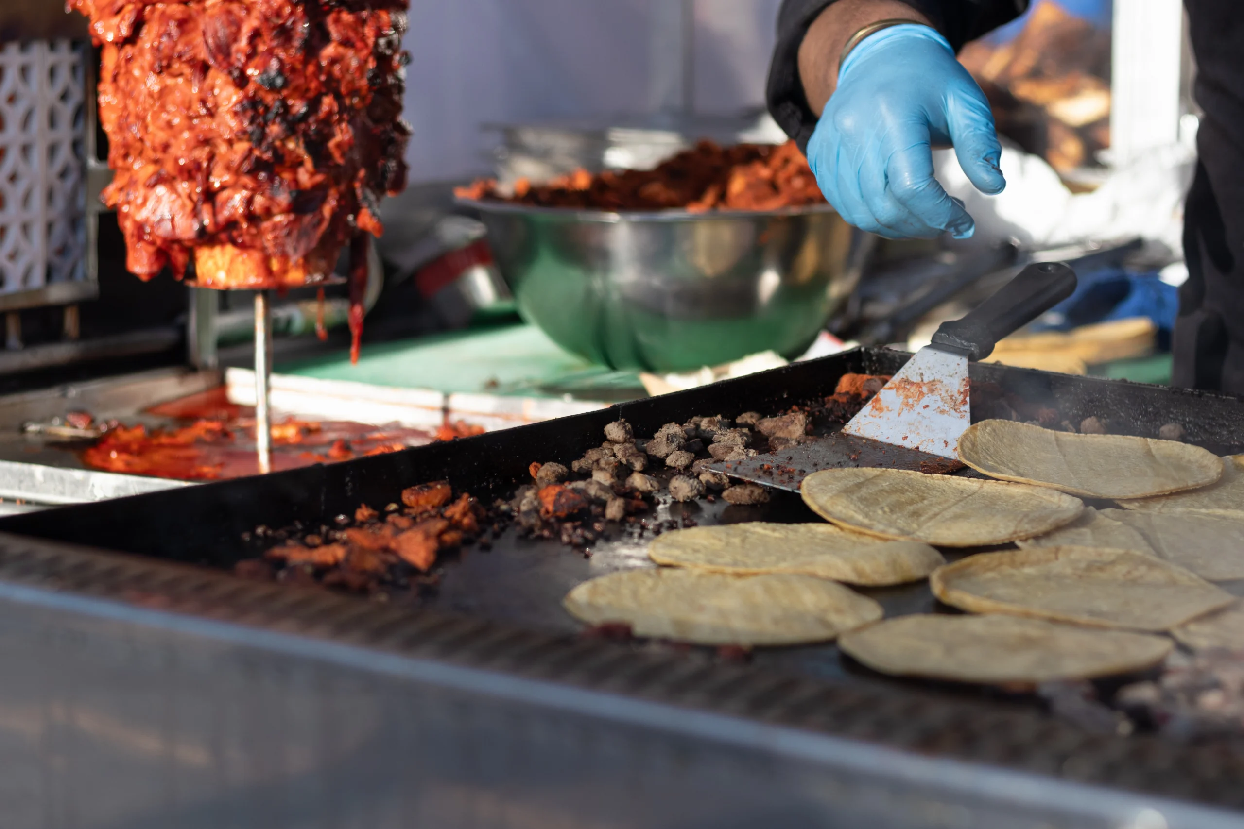 A taco stand griddle in daylight: a trompo of al pastor at one end, chopped meat and corn tortillas warming, and a gloved hand reaching for a spatula.